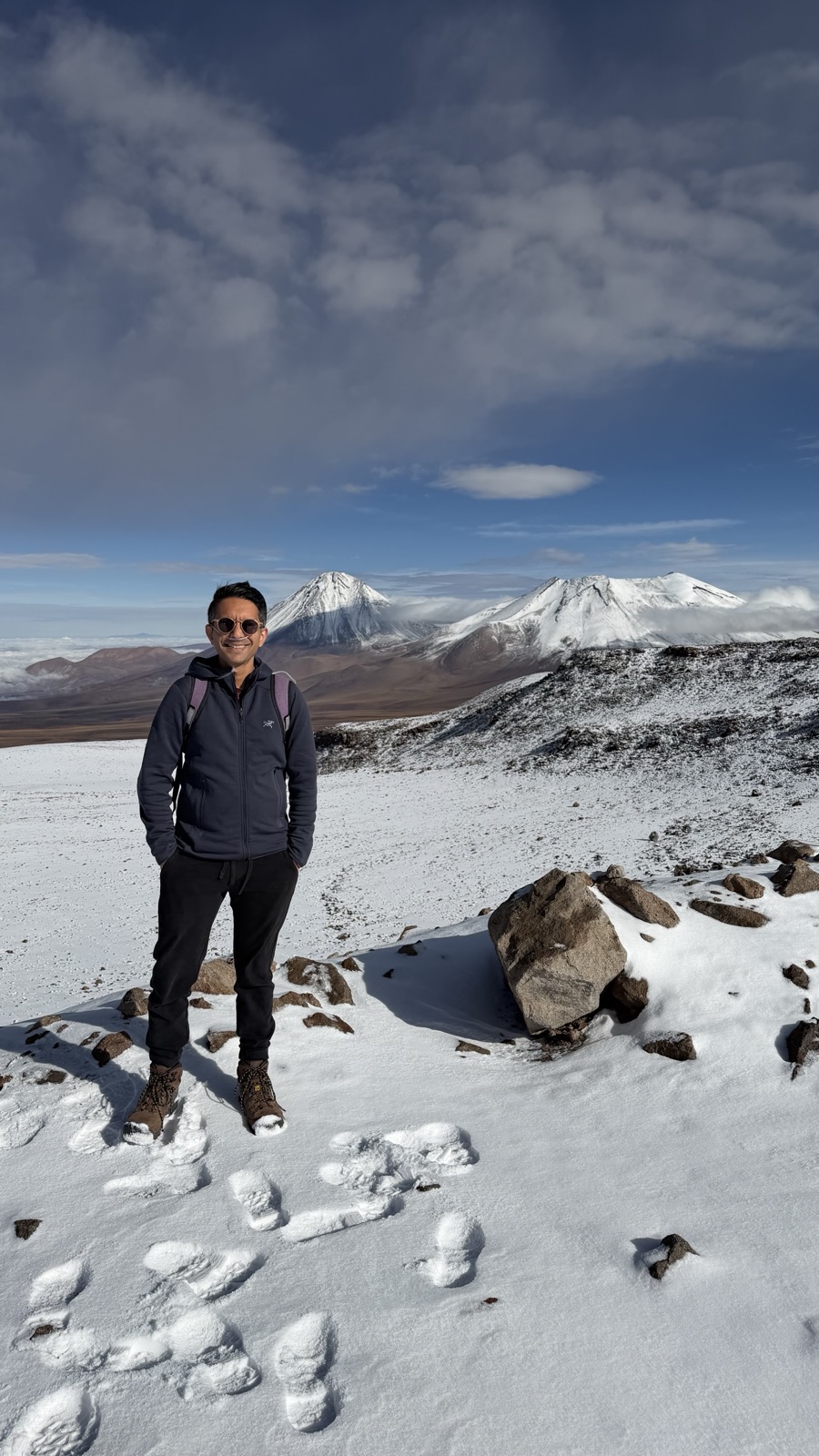 A person standing on a snow-dusted high-altitude plateau with volcanic peaks behind.
