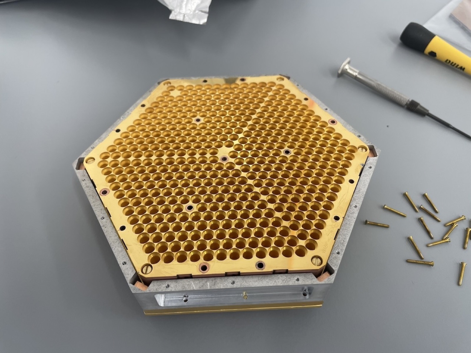 A gold hexagonal detector module on a bench, its face densely packed with circular feedhorn apertures, with assembly pins and a screwdriver beside it.