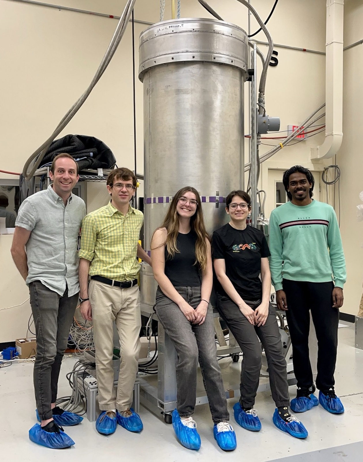 Five people standing beside a tall cylindrical cryostat in a laboratory high bay.
