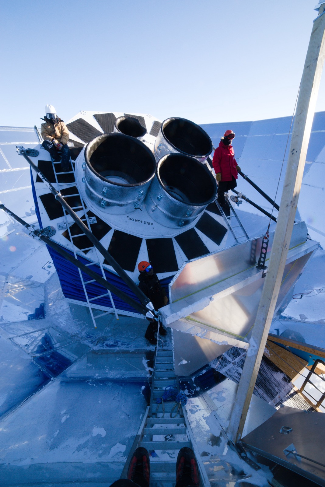 Two people on scaffolding beside the four cylindrical BICEP Array receivers, mounted in a blue-skirted ground shield on the Antarctic plateau.