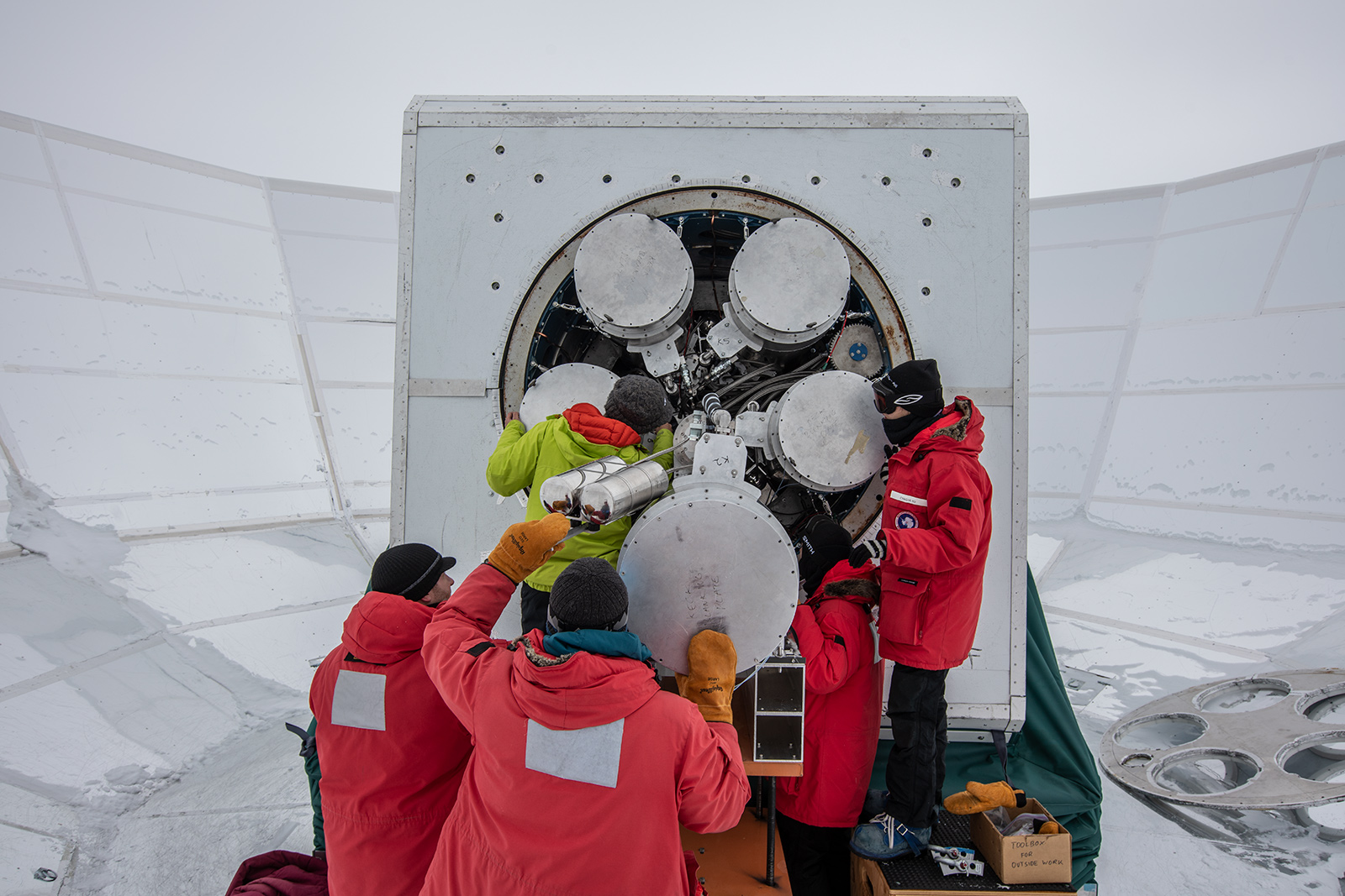 Several people in polar gear working on the open back of a telescope receiver mounted inside a large reflective ground shield, surrounded by snow.
