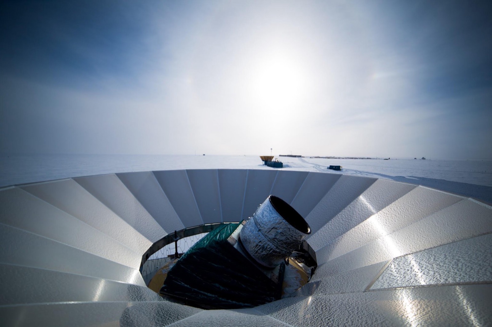 A telescope receiver pointing skyward from the center of a large white segmented ground shield, with the low sun and flat snow horizon behind it.