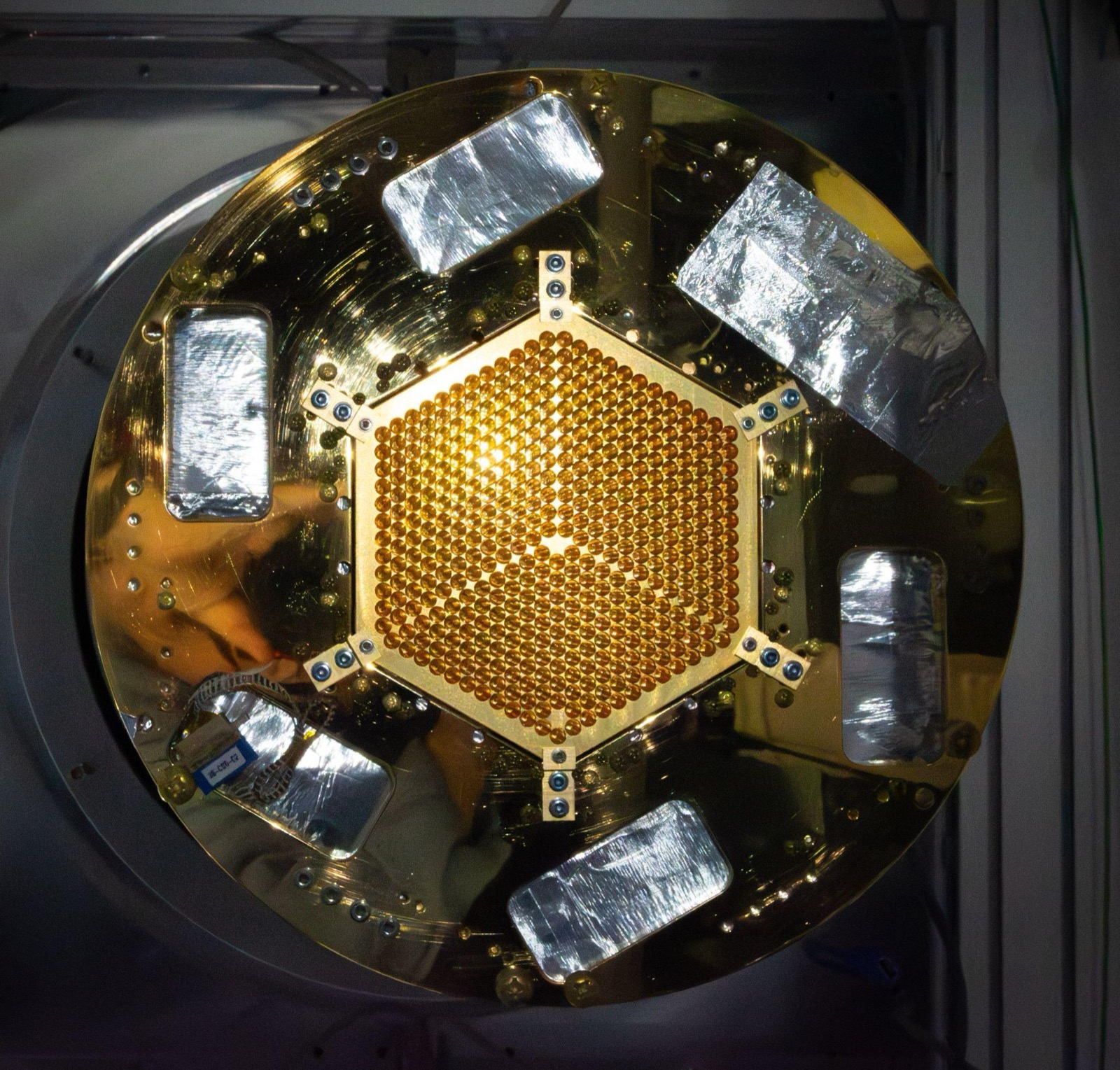 A gold-plated circular cryostat stage viewed head-on, with a hexagonal detector array of hundreds of small circular apertures at its center, surrounded by foil-wrapped components.