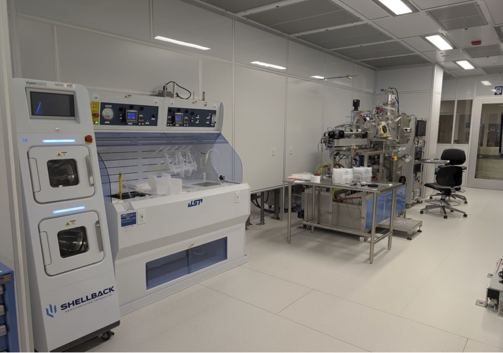 A cleanroom process bay with a wet processing tool in the foreground and a vacuum chamber system on the far wall.