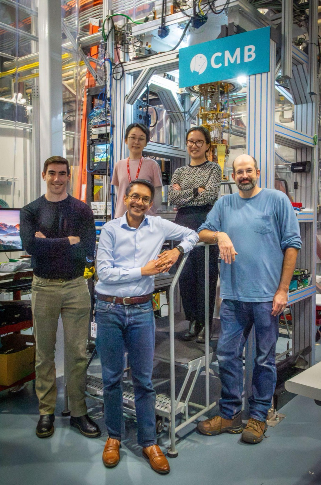 Five people standing and smiling in front of a laboratory cryostat rack with a sign reading CMB.