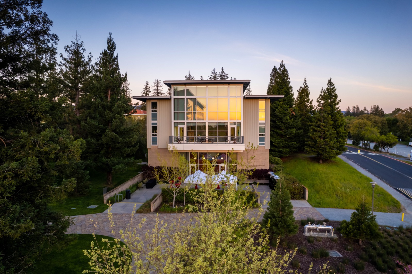 A modern three-story building with large windows, surrounded by redwoods and lawn at dusk.