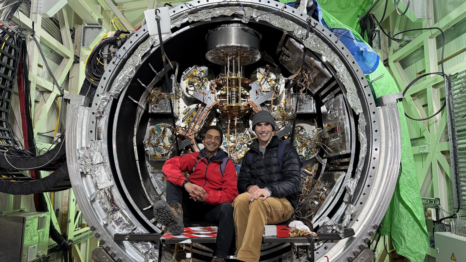 Two people seated in front of a large open cryostat packed with foil-wrapped optics tubes and wiring.