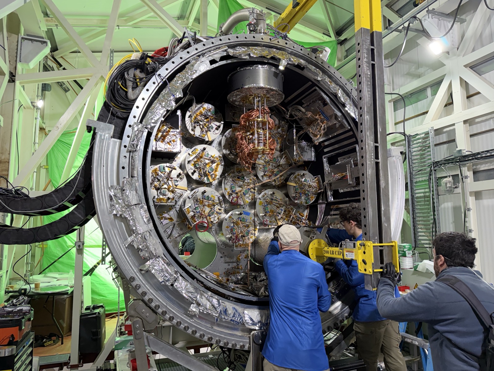 A large cylindrical cryostat opened to show a ring of foil-wrapped optics tubes, with people working at its rim.