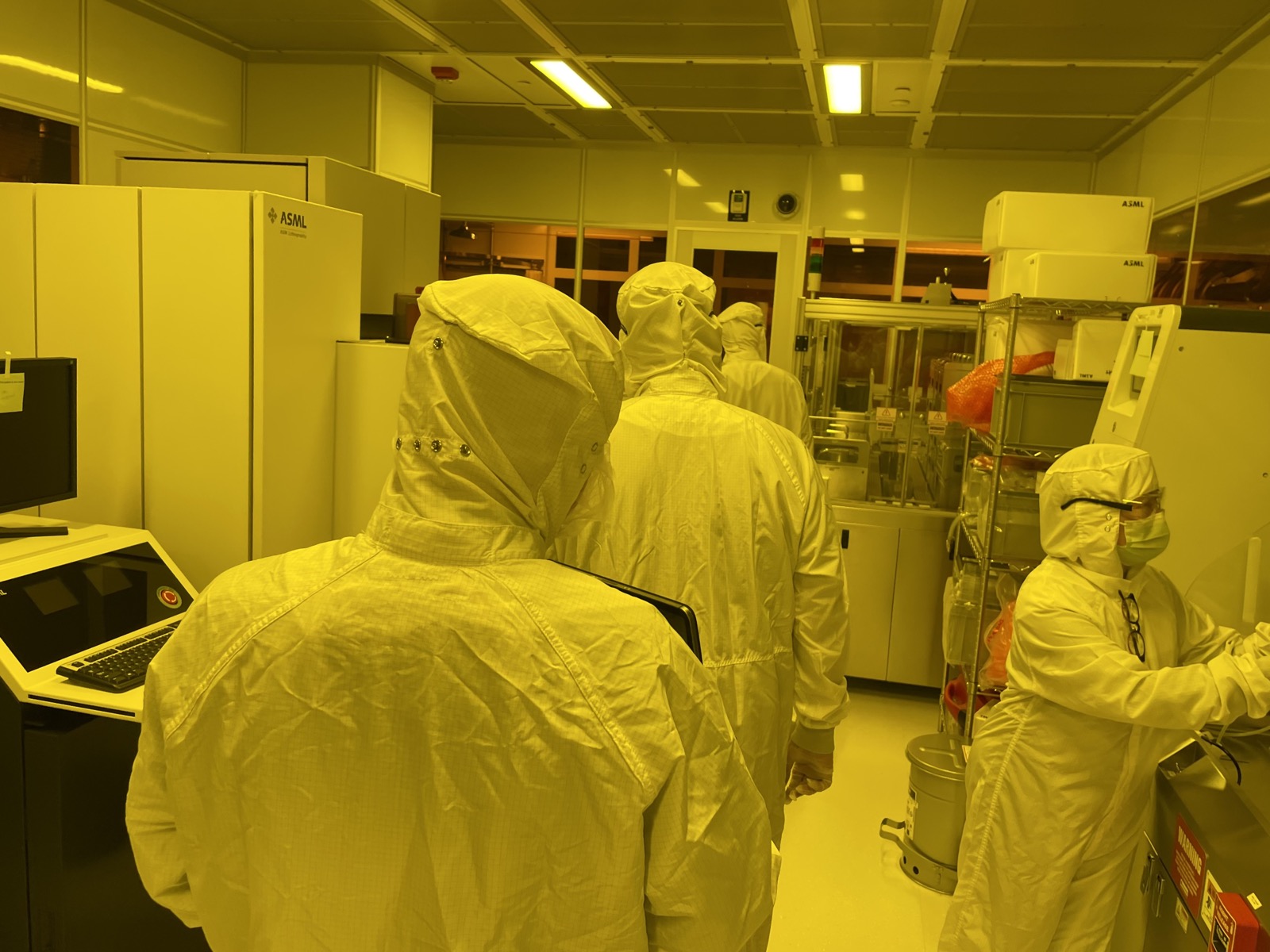 Four people in white cleanroom suits working in a bay lit entirely in yellow, beside a large lithography tool and racks of wafer carriers.