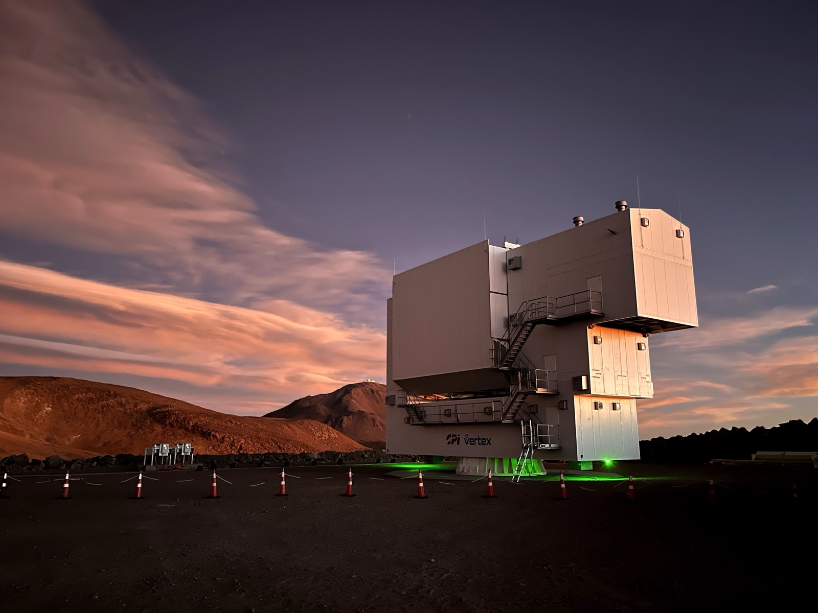 A large boxy telescope enclosure on the Atacama plateau at dusk, lit from below, with pink cloud and dark hills behind it.