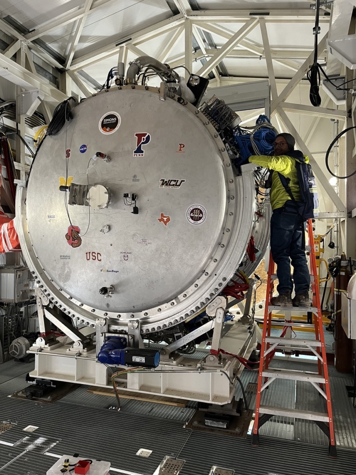 A person on a step ladder working at the side of a large cylindrical cryostat in a telescope enclosure.