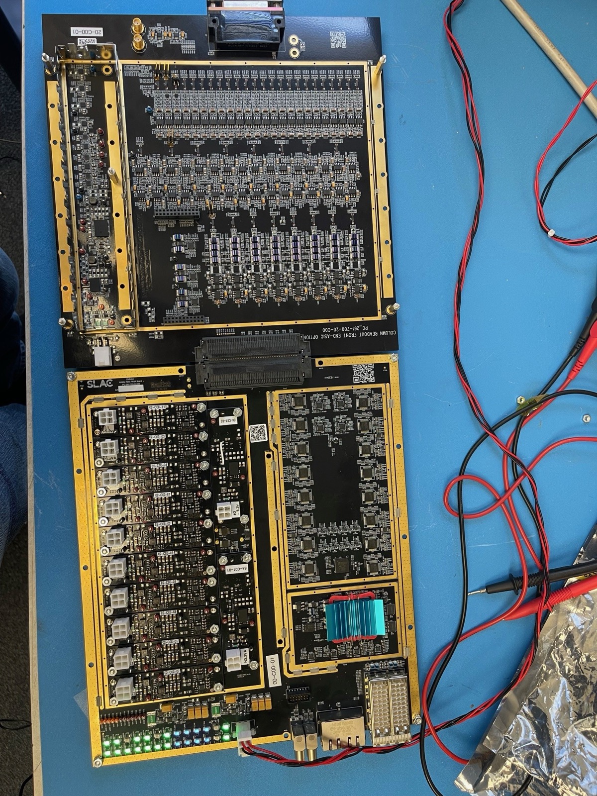 Two mated circuit boards on a bench, one carrying rows of gold-shielded modules and the other a dense array of components, with test leads attached.